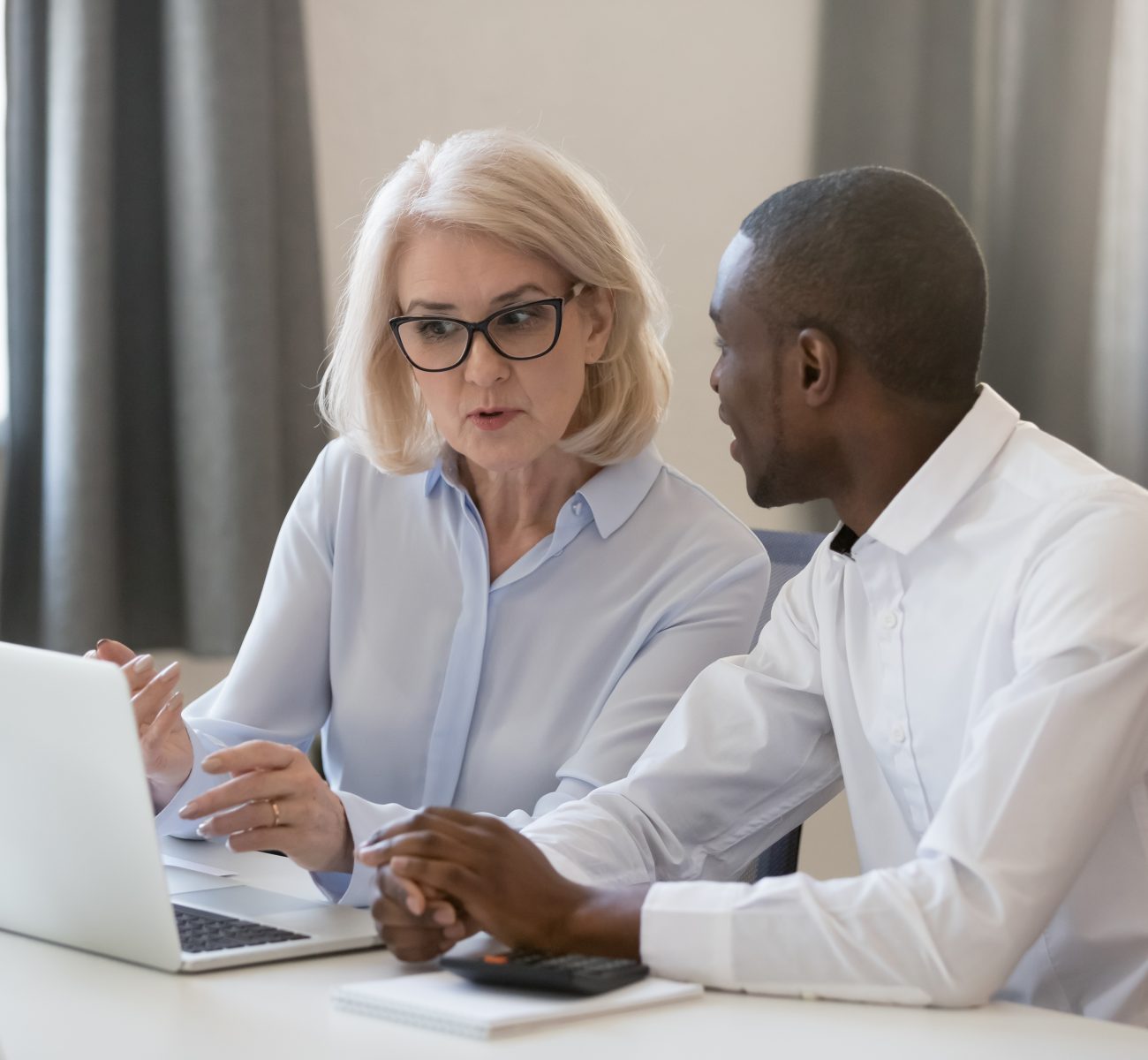 Serious experienced aged businesswoman in glasses explains help to african new employee task usage of corporate online program sitting at table office room working together, mentor and trainee concept